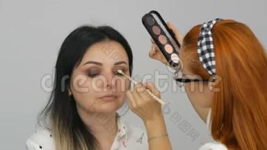 美丽的红发化妆师在一家美容院为一位年轻女子化妆，<strong>调色盘</strong>为米色眼影